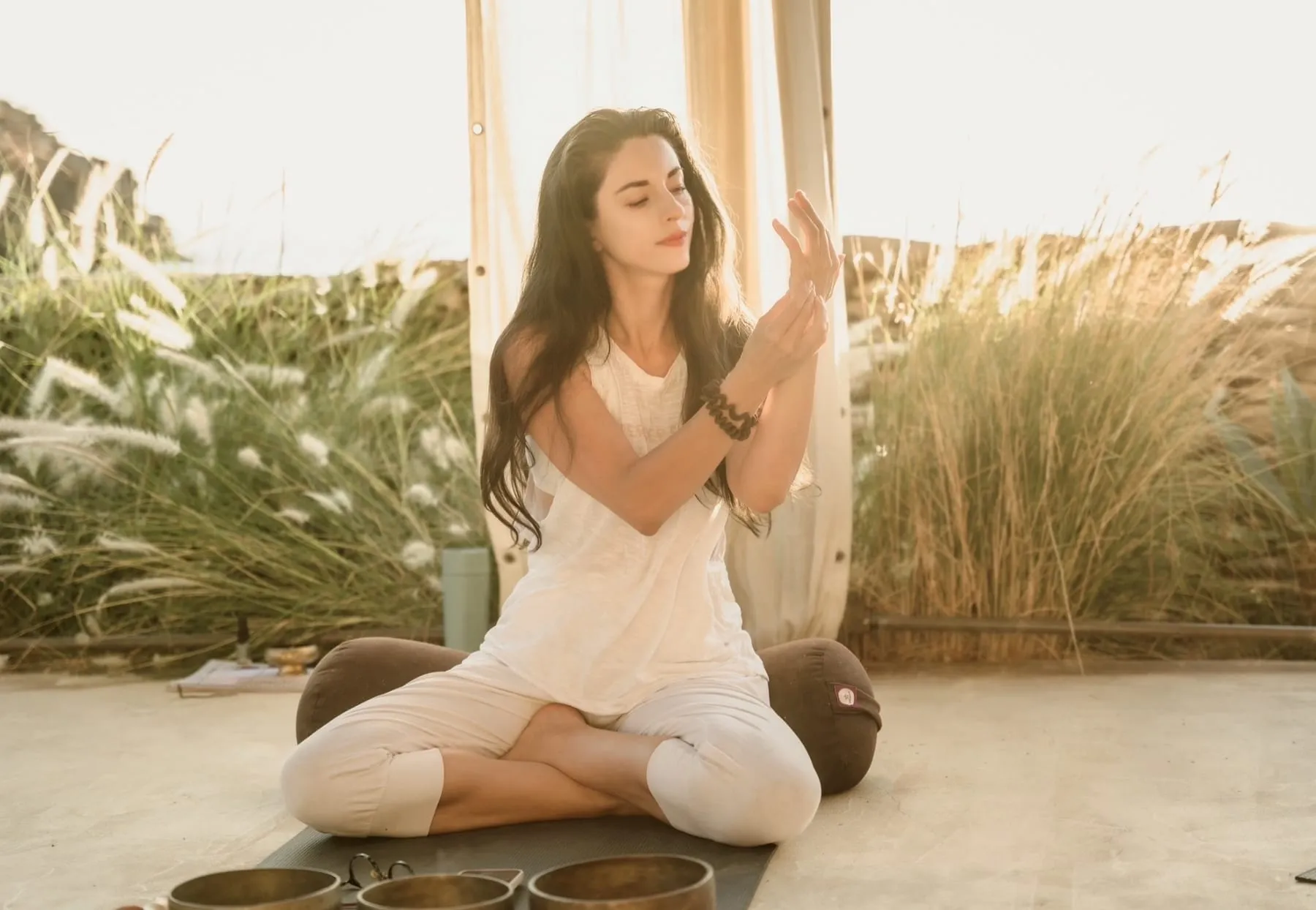 Dalia seated cross-legged in white linen, hands raised, singing bowls in the foreground